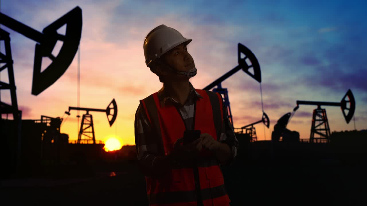 ingeniero masculino asiático con casco de seguridad usando un teléfono inteligente y mirando a su alrededor mientras está de pie frente a las bombas de aceite, durante la puesta o el amanecer