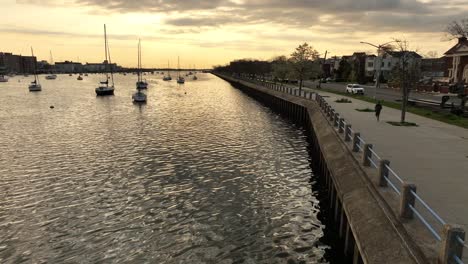 Eine-Aufnahme-Aus-Geringer-Höhe-über-Der-Sheepshead-Bay-Während-Eines-Goldenen-Sonnenaufgangs-Mit-Verankerten-Booten-Und-Einem-Jogger,-Der-Auf-Dem-Gepflasterten-Gehweg-Am-Wasser-Entlang-Läuft