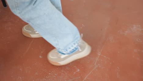 lower view of woman wearing blue shoes and jeans walking across reddish workshop floor towards electric sewing machine, places foot on stepper preparing to operate sewing machine