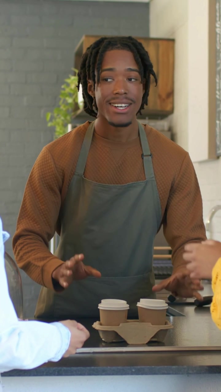 Vertical video: Bringing barista handing two paper cups after order ready at coffee bar, gray apron