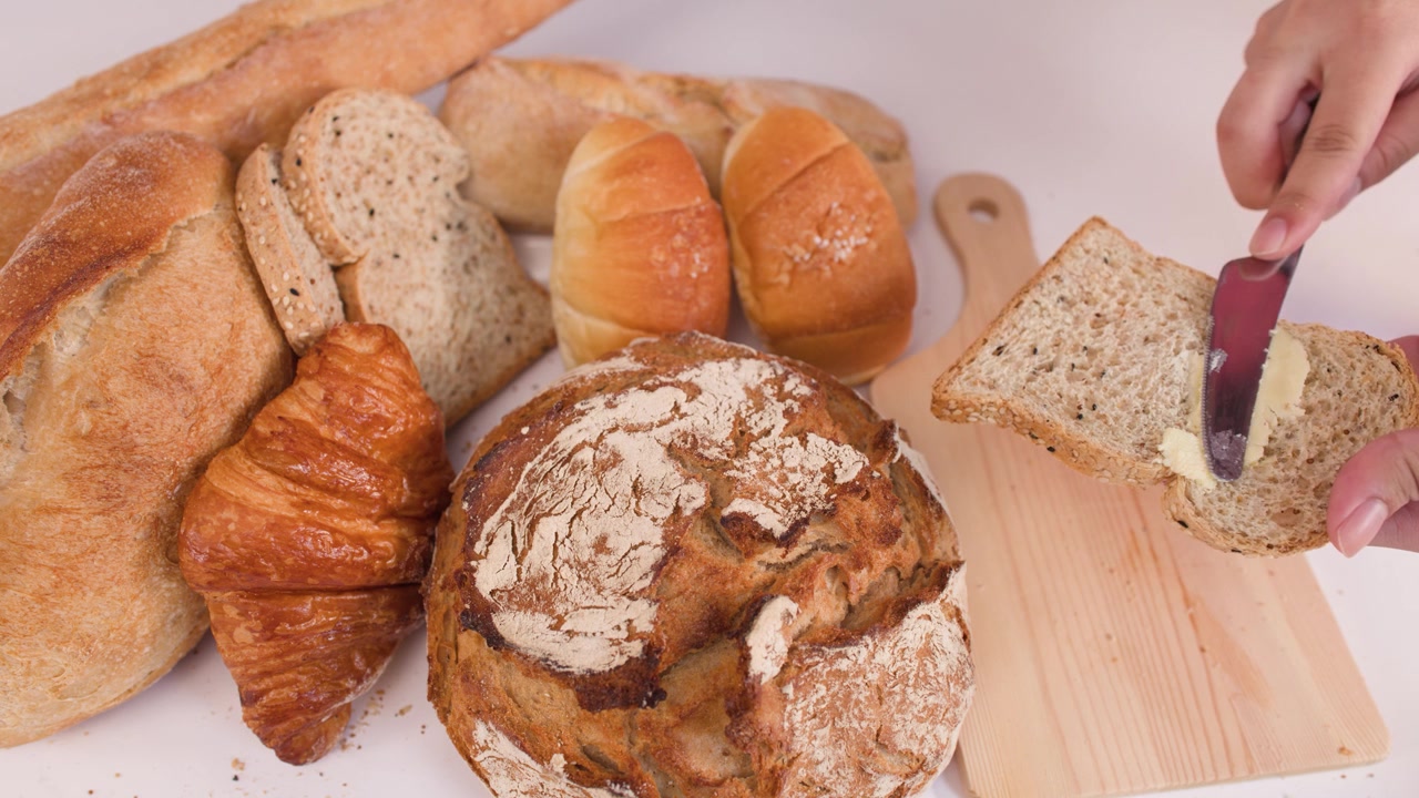 Close-up of hands spreading butter on multigrain bread slice amidst sourdough, croissants, and baguettes.