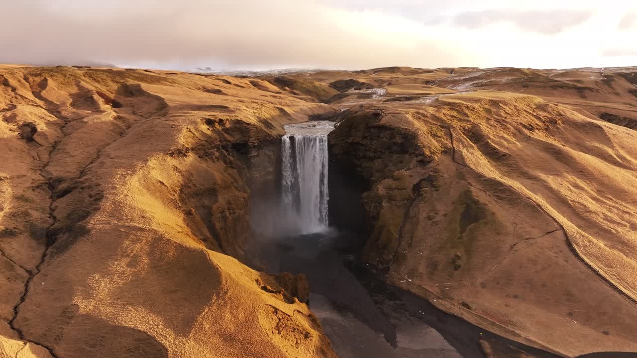 Aerial shot of Skógafoss waterfall in Iceland, surrounded by golden hills and dramatic valleys.