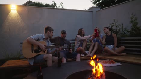 Happy-group-of-five-young-adults-chatting-and-having-fun-during-their-party-near-a-bonfire-in-the-evening-in-the-courtyard-of-a-country-house