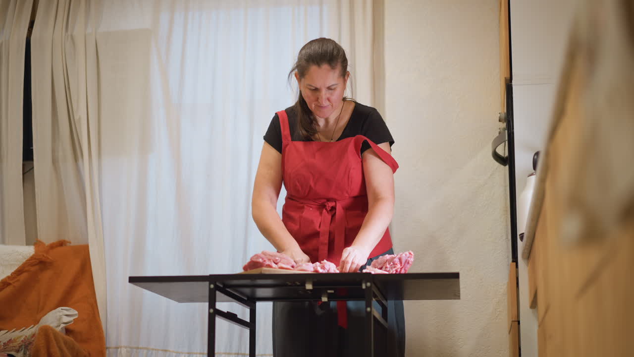 Kaukasische Hobbyköchin, ungestellt aufgenommen. Vordergrundunschärfe mit weichen, warmen Tönen. Rote Schürze, Hände arrangieren Zutaten auf dem Tisch. Ein langsames häusliches Ritual, gemütliche Essecke mit natürlichem Fensterlicht.