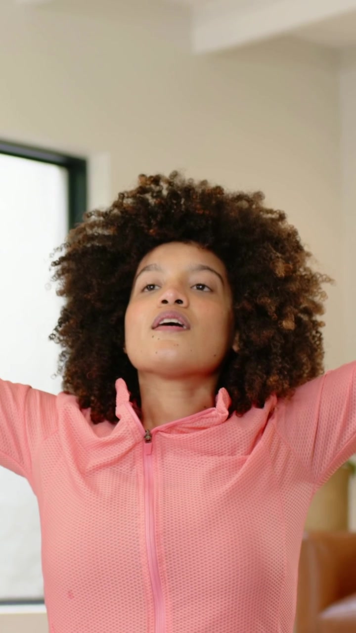 Vertical video: Breathing woman in pink jacket, light leggings stretching on purple mat at home