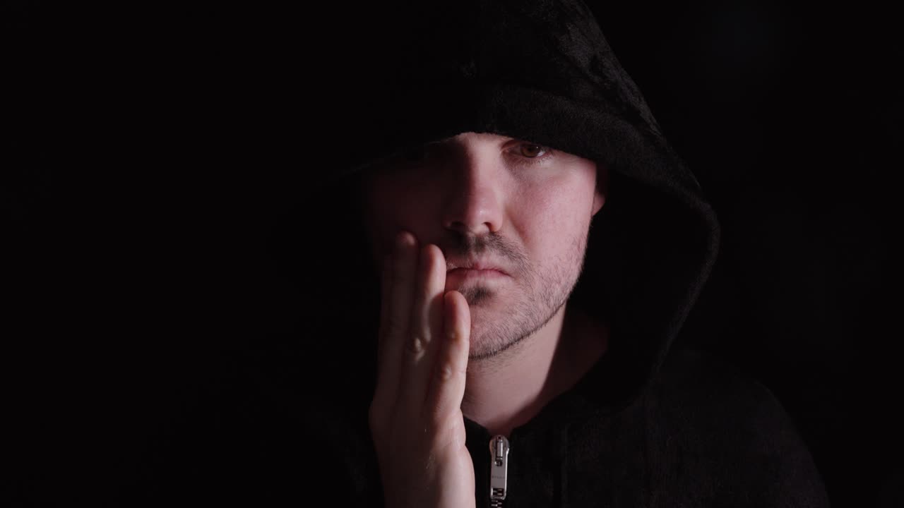 Uomo con felpa scura con cappuccio che pensa con la mano sul mento, a simboleggiare mistero, contemplazione e pensieri interiori.