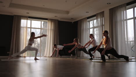 Club-De-Fitness.-Instructora-Caucásica-Entrenando-Y-Ajustando-La-Pose-Correcta-A-La-Estudiante-Caucásica-Al-Frente-Mientras-Otros-Las-Siguen.