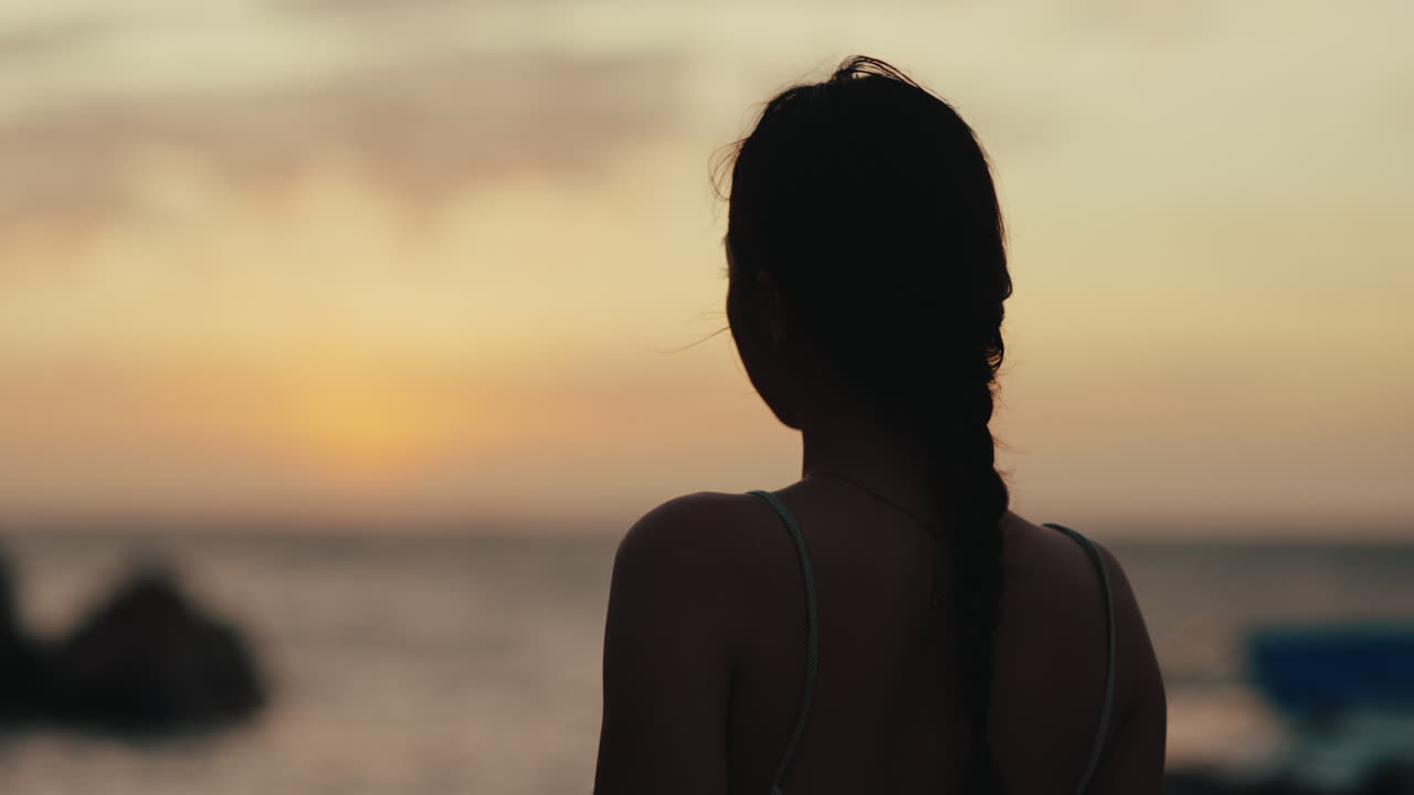 年輕女子在岩石海岸線邊觀看平靜海面上的日落剪影，享受寧靜祥和的獨處時光。