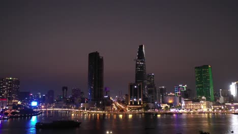 Ho-Chi-Minh-City,-Vietnam-iconic-Skyline-and-Saigon-river-waterfront-aerial-panorama-on-a-busy-evening-featuring-all-key-buildings-illuminated-with-a-container-boat-in-silhouette-on-the-river
