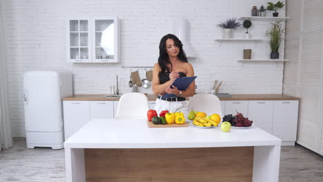 Attractive woman in kitchen choosing products for a healthy diet. Female taking notes about fruit and vegetables on the table in front of her.