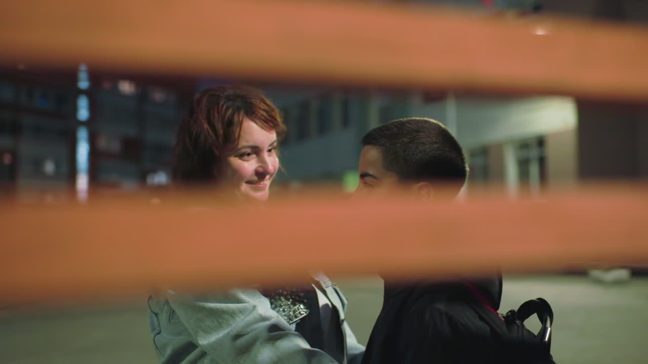 Mother gently adjusts boy’s jacket with smile during early morning outside school, viewed through wooden bench slats, capturing warm emotional moment of care, protection, and daily routine before school begins