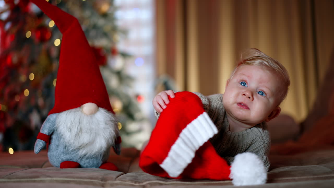 Adorable blond baby boy wearing warm sweater lies near the toy gnome. Kid looks up holding a Santa cap in hands. Blurred Christmas tree at backdrop.