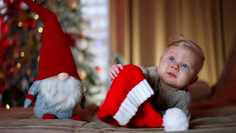 adorable blond baby boy wearing warm sweater lies near the toy gnome. kid looks up holding a santa cap in hands. blurred christmas tree at backdrop.
