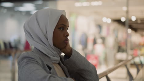 unhappy muslim woman in hijab leans on handrail in mall