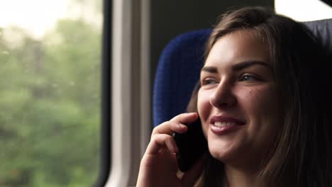 Nahaufnahme-Des-Gesichts-Der-Frau.-Hübsches-Mädchen-Fährt-Mit-Dem-Zug.-Telefoniert-Mit-Ihrem-Handy.-Lächelnd,-Entspannt.-Bewegtes-Naturbild-Im-Schaufenster