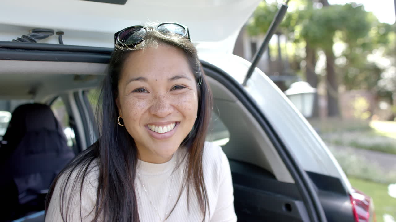 mujer asiática feliz con gafas de sol sentada en el maletero del coche y sonriendo en un día soleado, en cámara lenta