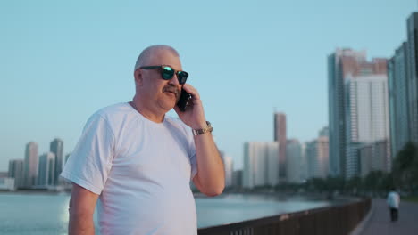 happy handsome senior man in sunglasses and white t-shirt talking on the phone standing on the waterfront in summer against the city and buildings