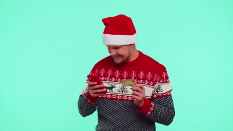 Man-in-Christmas-red-sweater-use-mobile-cell-phone-and-plastic-credit-bank-card,-win,-calebrate,-wow