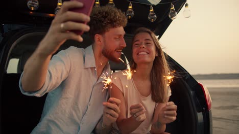 A-bearded-man-with-curly-hair-in-a-blue-shirt-and-his-blonde-girlfriend-are-holding-Bengal-lights,-smiling-and-taking-photos-on-the-phone-in-the-red-phonecase-in-the-trunk-of-the-black-car-decorated-with-the-lights-against-the-background-of-the-yellow-sky