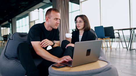positive brunette holding a cup talks cheerfully to a man. male colleague is focused on laptop standing in front of him. cooperation and communication concept.