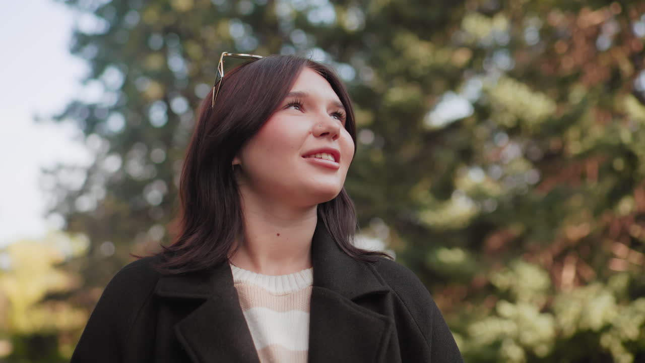 가을 캐노피 아래에서 다크 코트와 스트라이프 스웨터를 입은 패셔너블한 소녀가 따뜻한 미소로 햇살 가득한 공원의 자연을 감상하며 위를 올려다보고 있어요. 뒤로는 부드러운 보케 나무가 드리워져 있어 즐거운 야외 순간을 연출하네요.