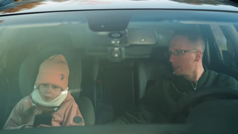 a thoughtful moment captured inside a car, as a man in a green jacket attentively explains something to a little girl in a pink beanie and jacket, who is focused on a smartphone in her hands