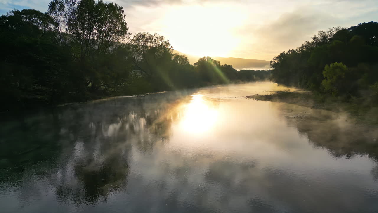 Aerial drone view of a river at sunrise with morning mist floating above the water and golden sunlight reflecting on the surface in Baja
