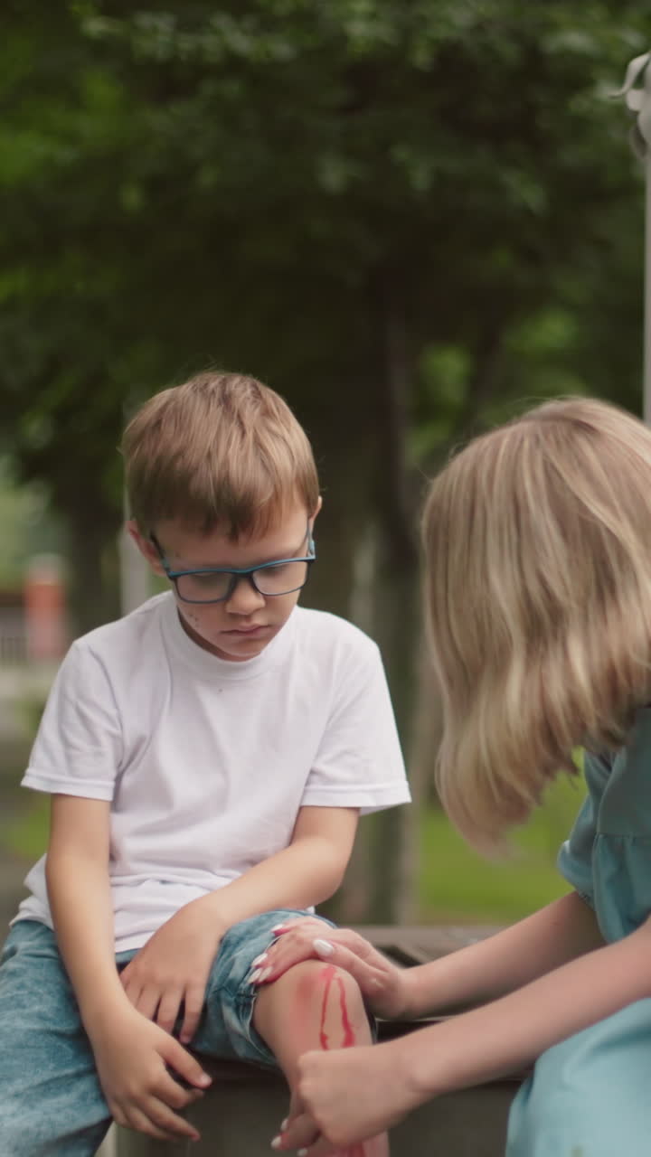 una madre se arrodilla al lado de su hijo en una tranquila carretera del parque, limpiando suavemente su herida en la pierna mientras él mira hacia abajo, visiblemente triste