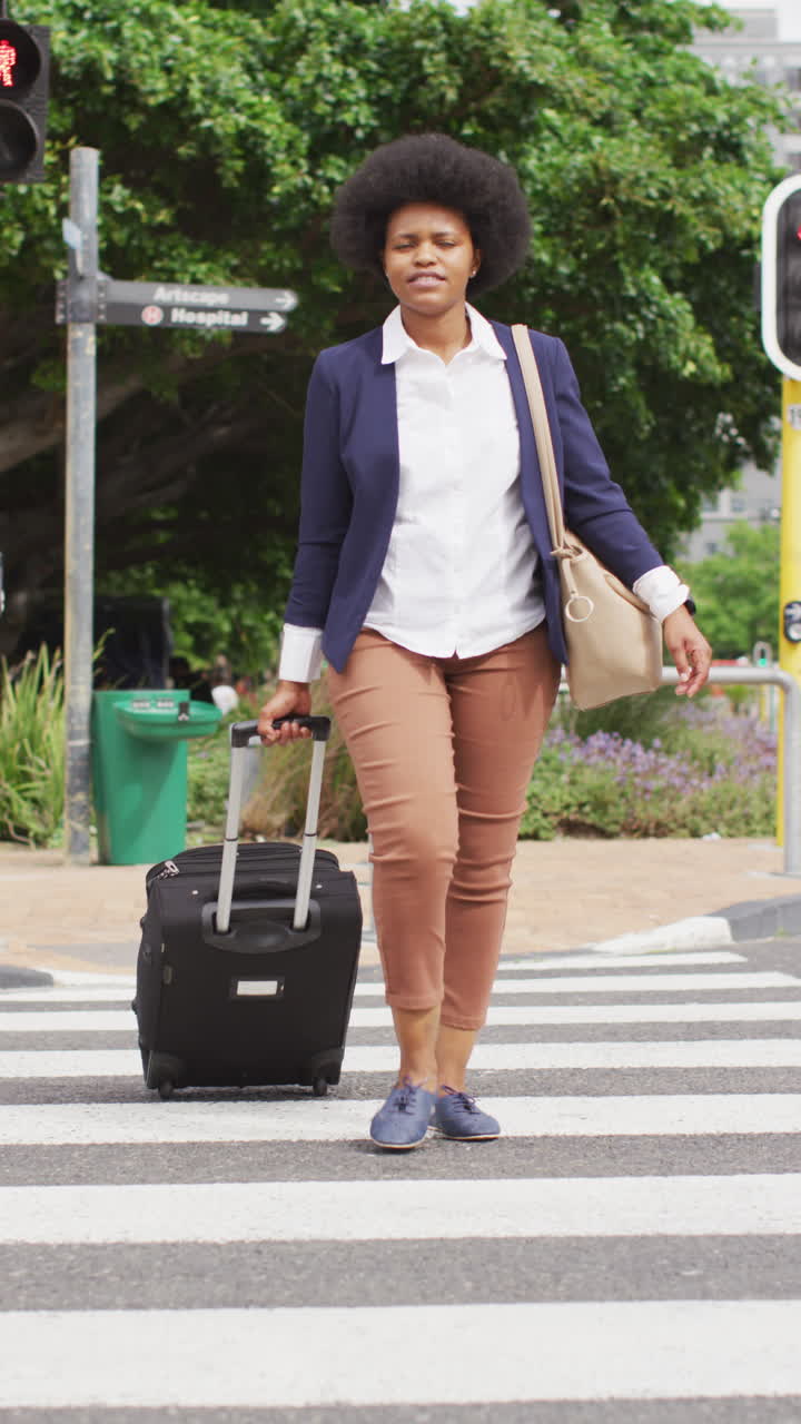 video vertical de una feliz mujer afroamericana caminando en la ciudad con una maleta de ruedas, mirando a su alrededor