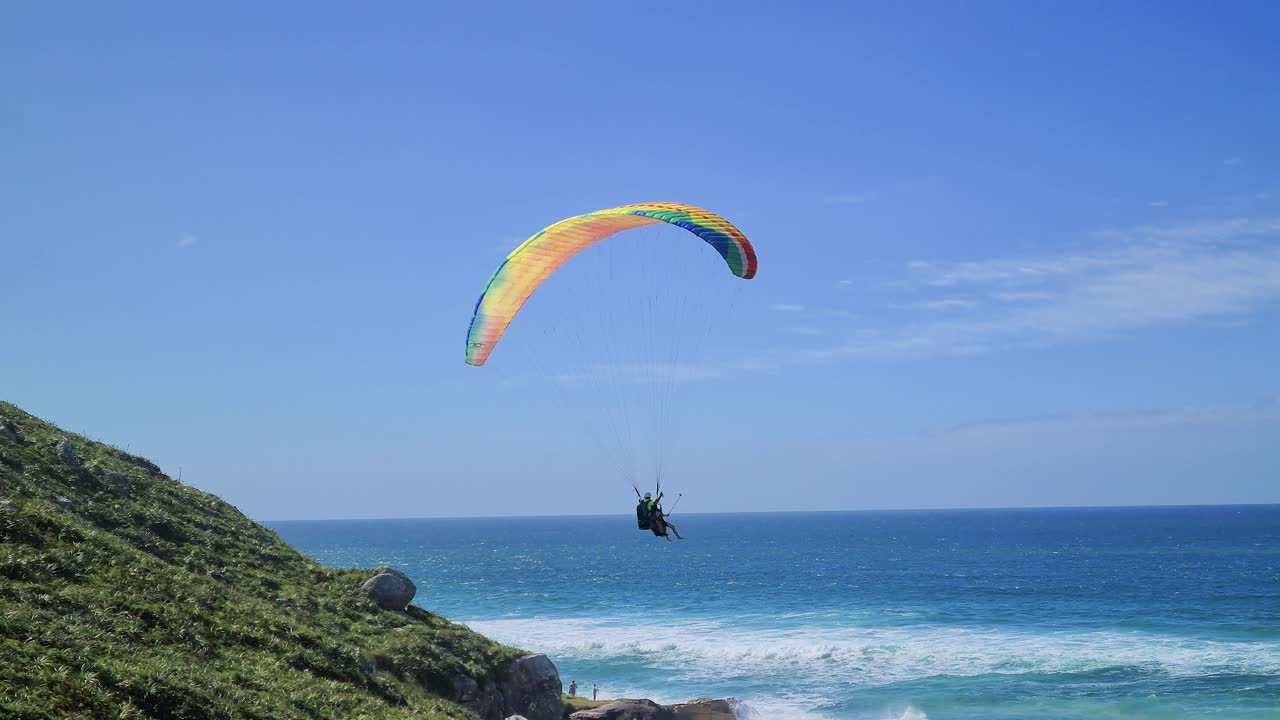 escena de vuelo en parapente frente a la playa con rocas, montañas y arena con gente divirtiéndose en el vuelo con olas marinas deporte aéreo