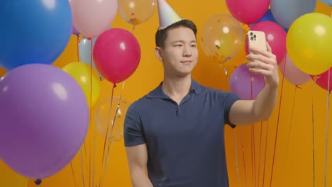 studio portrait of man taking selfie wearing party hat celebrating birthday surrounded by balloons