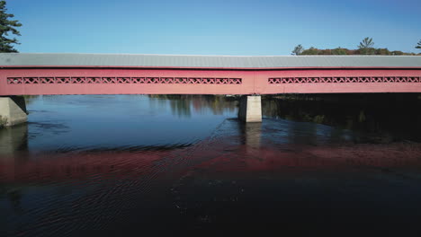 an aerial drone view underneath a red and gray covered walking bridge across a river during the fall season