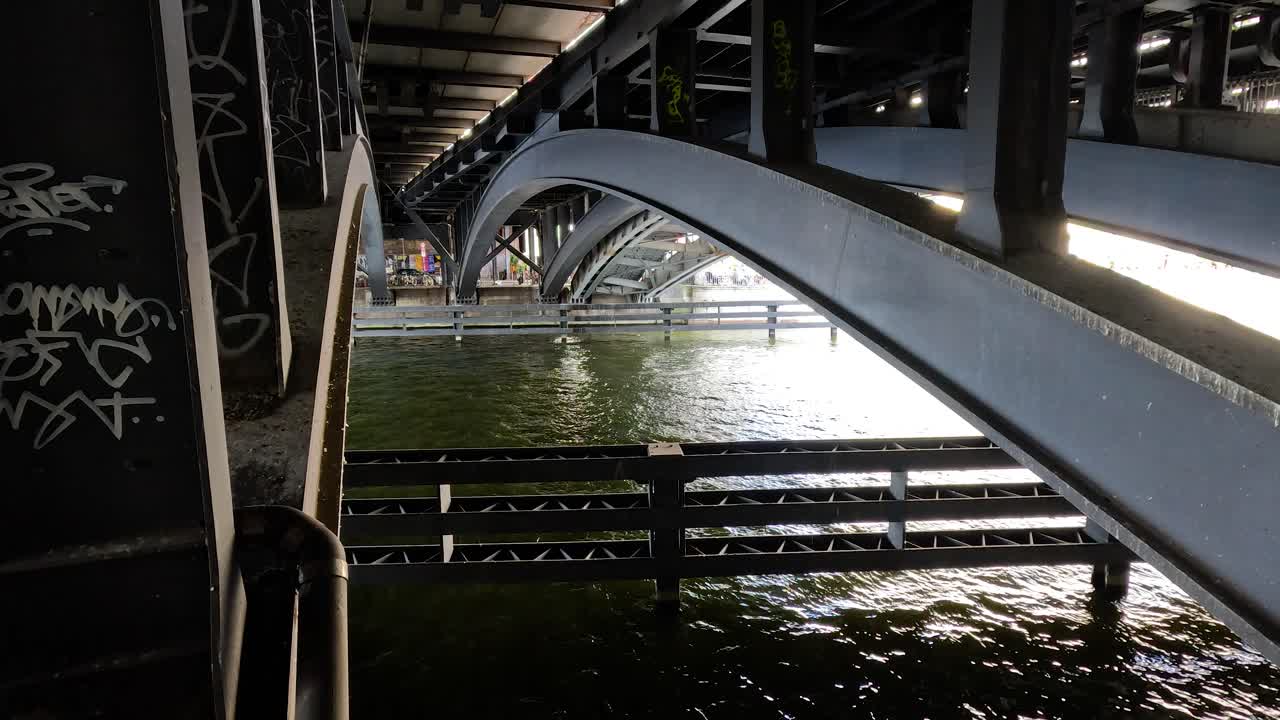 Steel bridge arches span above a flowing river in an urban setting, with graffiti on support columns. Natural daylight, steady camera, moody atmosphere