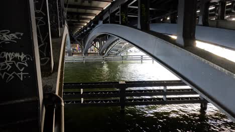 steel bridge arches span above a flowing river in an urban setting, with graffiti on support columns. natural daylight, steady camera, moody atmosphere