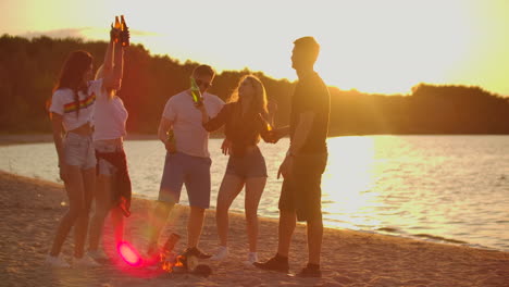 Drei-Mädchen-Und-Zwei-Jungen-Verbringen-Einen-Sommerabend-Bei-Sonnenuntergang-In-Shorts-Und-T-Shirts-Am-Strand-Mit-Bier-Am-Lagerfeuer.-Sie-Stoßen-Mit-Bier-An-Und-Tanzen-Auf-Der-Open-Air-Party.