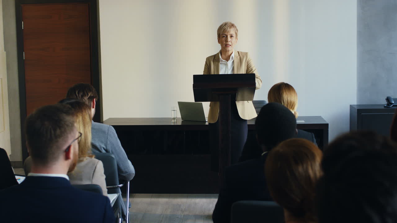 Caucasian businesswoman wearing formal clothes and talking on a podium in a conference room in front to many people