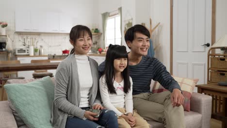 beautiful smiling asian family of three sitting together watching tv at home. mom tuning channels with controller and dad explaining to their daughter with finger pointing