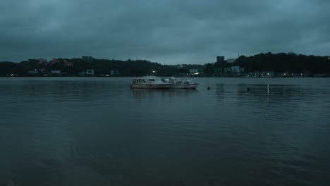 Boot-Vor-Anker-Auf-Einem-Ruhigen-See-In-Der-Abenddämmerung-Mit-Einer-Skyline-Der-Stadt-Im-Hintergrund