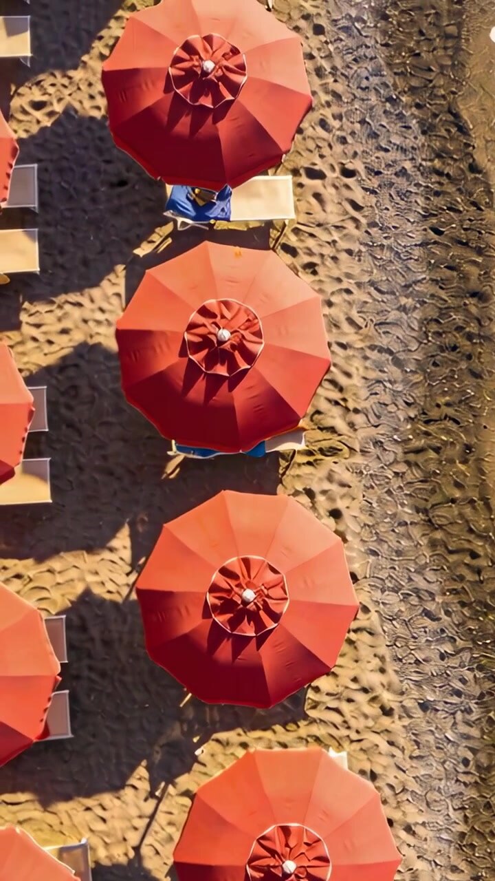Video verticale: Pilota che avvia la discesa del drone, telecamera che scivola lungo la spiaggia, rivelando ombrelloni rossi