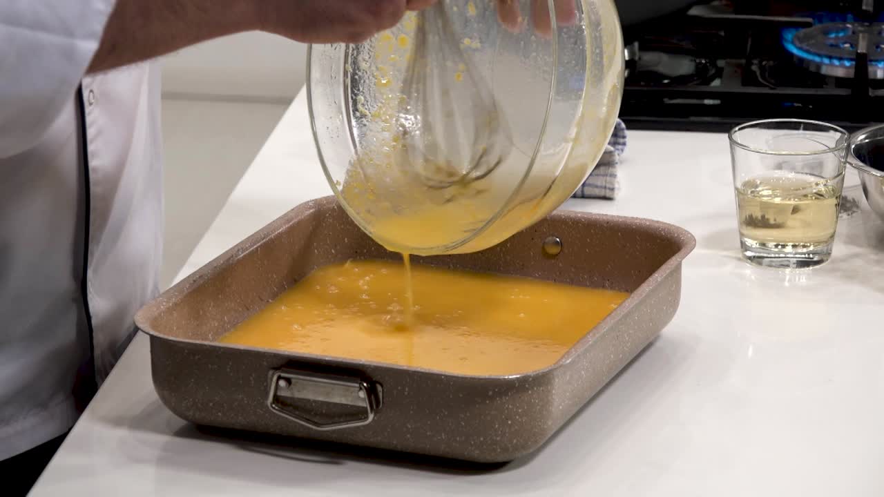 A chef skillfully whisks and pours a creamy egg mixture into a baking dish. The contemporary kitchen is well-equipped and bright, creating a vibrant cooking atmosphere