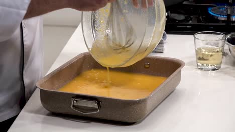 a chef skillfully whisks and pours a creamy egg mixture into a baking dish. the contemporary kitchen is well-equipped and bright, creating a vibrant cooking atmosphere