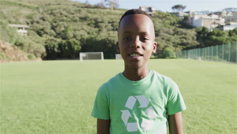 An-African-American-boy-wearing-a-green-recycling-t-shirt-stands-on-a-sunny-soccer-field,-with-copy-