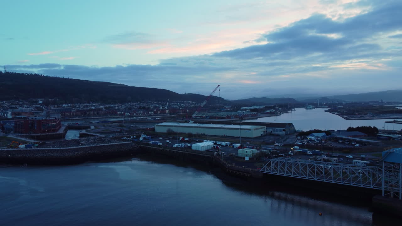 Umkreisende Luftaufnahme der Swansea Docks bei Sonnenaufgang im Blaue-Stunde-Licht. Maritime Industrielandschaft zum Anlegen von Frachtschiffen sowie zur Bootsreparatur und -wartung.