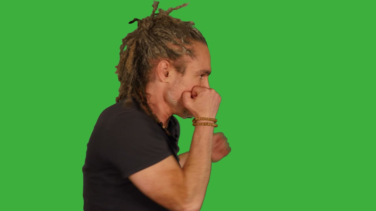 Middelaldrende mann kjemper foran en grønnskjerm. Forbereder seg på kamp og slår mot kameraet. Dreadlocks og svart T-skjorte.