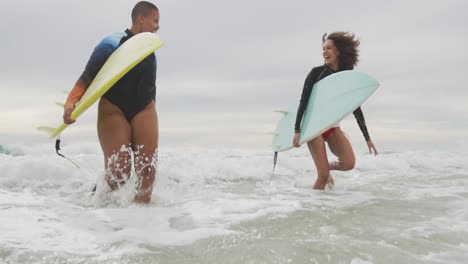 happy african american female friends running out of sea holding surfboards