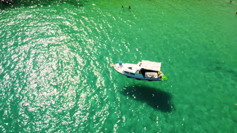 Bote-Pequeño-Flotando-En-Las-Aguas-Iluminadas-Por-El-Sol-Del-Océano-Con-Gente-Nadando-En-Un-Día-Caluroso-En-Verano-En-La-Playa-De-Prapratno,-Croacia