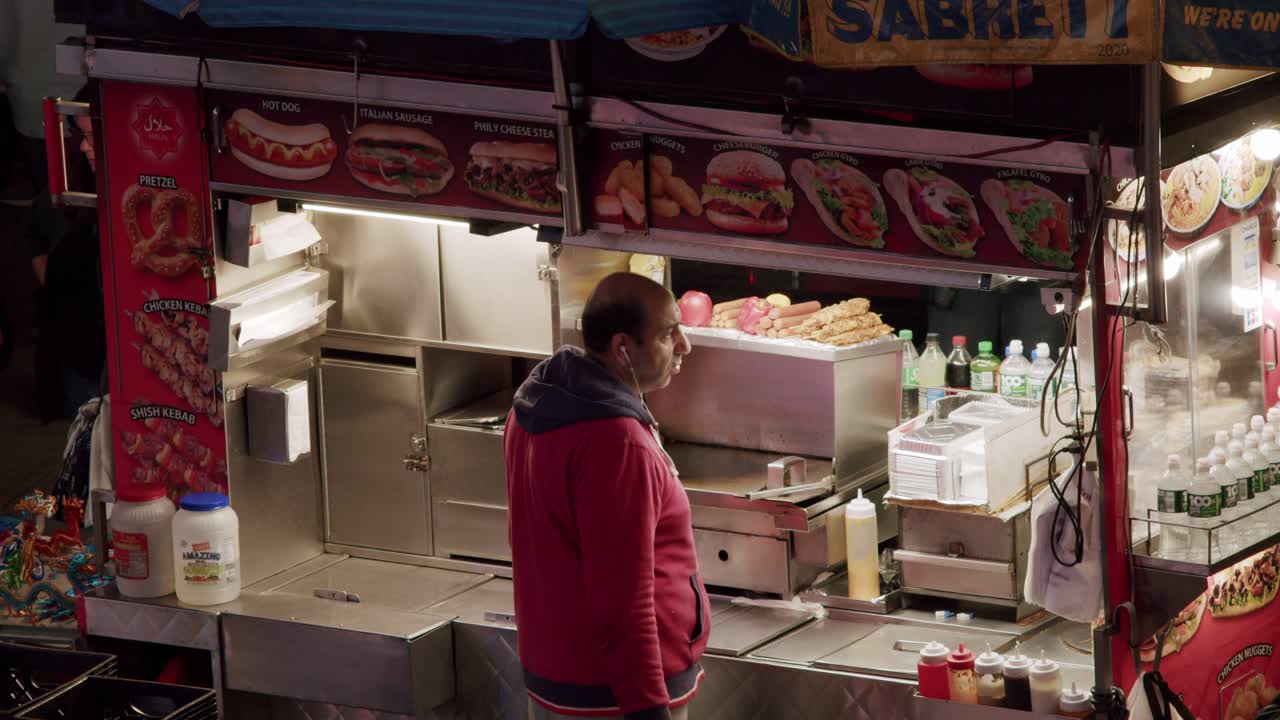 Man prepares food at a New York City street stand with colorful signs and hot dog options