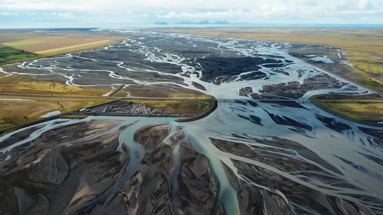 Aerial view capturing a glacial river winding through Icelandic plains, forming intricate patterns of water and sediment against a backdrop of mountains and clouds, near Seljalandsfoss waterfall