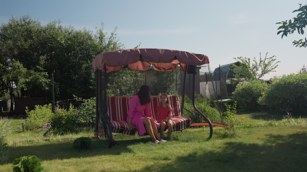 Mother And Young Daughter Sit Calmly On Colorful Outdoor Swing Surrounded By Lush Greenery, Woman And Her Child Rest Quietly On Striped Swing Beneath Canopy In Peaceful Garden Setting