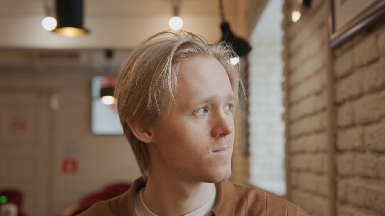 Close up of young man drinking from pink mug in indoor setting with soft lighting. subject is wearing brown shirt and background features blurred interior elements like hanging lights